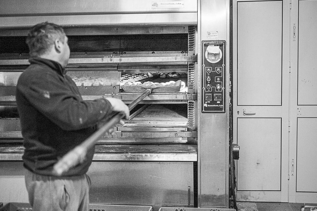 Rui Carvalho takes the baked bread out of the oven. He tells me about other more industrialized bread factories where in one hour is produced more than 10 times of his daily production. Rui Carvalho retira uma fornada de pão do forno. Ele conta-me histórias de fábricas de pão onde numa hora se produz mais de 10 vezes do que ele produz num dia inteiro.