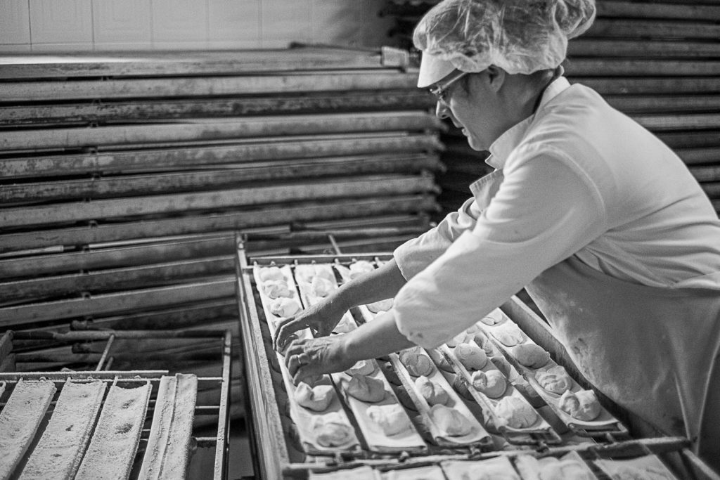 Márcia Carvalho places the previously shaped individual portions of dough on trays which will later carry them into the oven. These trays will simultaneously all turn around by means of a lever and make the dough fall on the oven surface. The trays are then removed out of the oven. Márcia Carvalho coloca as doses previamente modeladas em tabuleiros que posteriormente as levarão para o forno. Estes tabuleiros possuem um sistema de alavanca que faz virar e cair simultaneamente todas as doses na superfície do forno. O tabuleiros são depois removidos do forno.
